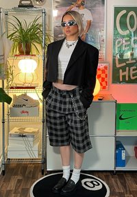 Črn skrajšan blazer preko belega majice, v kombinaciji s črno-belimi kratkimi hlačami s karirastim vzorcem, ki vključujejo verige. Črne čevlje in bele nogavice dopolnjujejo videz.