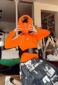 Oranssi käsilaukku, jossa on kierretty kahva ja käärmeviimeistely, yhdistetty mustaan vyöhön. Asusteeseen kuuluu vetoketjulla varustettu oranssi takki ja kuvioitu hame.