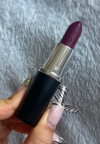MAC Lippenstift in einer Stiftform; tiefes Pflaumenrot; Metallgehäuse mit silbernem Band; schwarzer Sockel; glatte Textur; gehalten zwischen dekorierten Nägeln.