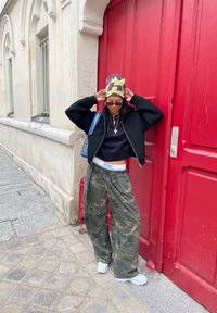 Veste noire zippée recadrée sur un haut noir, pantalon baggy camouflage et bonnet à motif. Debout à côté d'une porte rouge, trottoir gris.