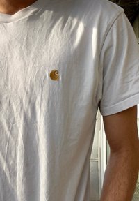 T-shirt de algodão branco com mangas curtas, apresentando um pequeno logótipo bordado dourado no lado esquerdo do peito. Textura suave, ajuste casual.