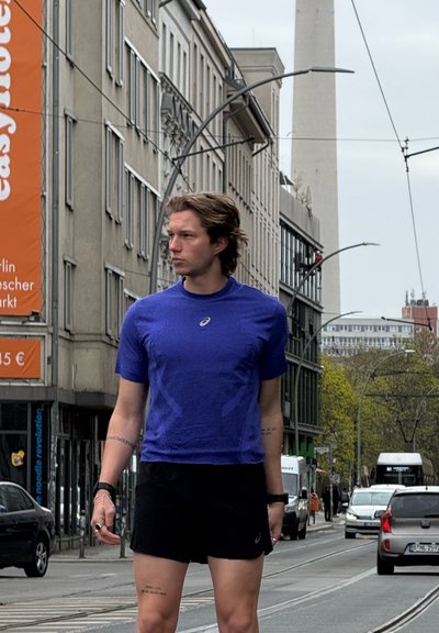 Joven con camisa azul y pantalones cortos negros de pie en una calle de la ciudad con coches y edificios, mirando hacia la izquierda, cielo nublado.