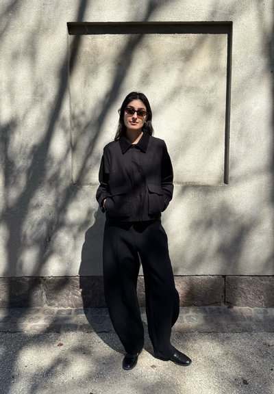 Mujer con gafas de sol oscuras, chaqueta negra y pantalones anchos negros está de pie con las manos en los bolsillos frente a una pared texturizada iluminada por el sol con sombras.