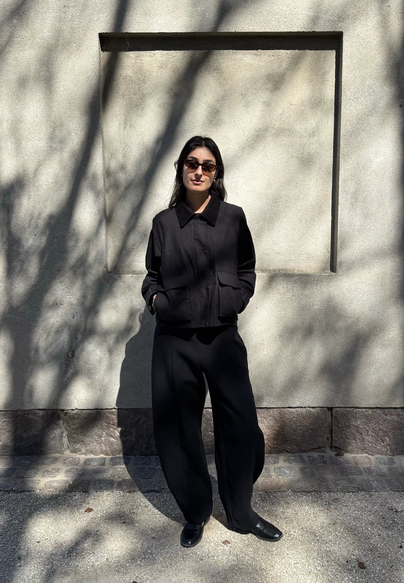 Femme portant des lunettes de soleil foncées, une veste noire et un large pantalon noir se tient les mains dans les poches devant un mur texturé éclairé par le soleil avec des ombres.