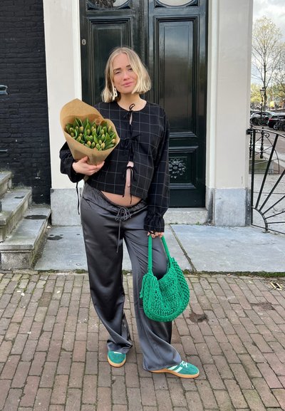 Top a cuadros negros con lazos, pantalones de satén grises, bolso tejido verde y zapatos verdes. Sosteniendo un ramo de tulipanes envuelto en papel kraft.