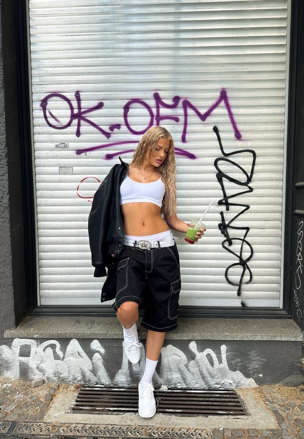 Calças cargo pretas com costuras brancas, top cropped branco, casaco oversized preto e ténis brancos. Segurando uma bebida verde. O fundo apresenta graffiti.