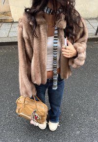 Personne portant une veste en fourrure marron, un crop top blanc, un jean bleu, une écharpe rayée, des chaussures crème, tenant un sac à main beige avec des accessoires porte-clés.