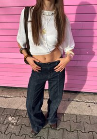White cropped jumper with puff sleeves, dark wide-leg denim jeans, layered necklaces, and bracelets. Background features a pink wall.