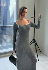 Szürke és fehér csíkozott, hosszú ujjú ruha, négyzet alakú dekoltázzsal és testhez simuló fazonnal. A modell egy fekete táskát tart egy minimalista háttér előtt.