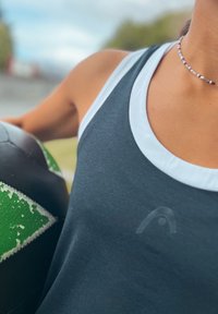 Sport tanktop mit einem dunkelgrauen Körper, weißem Kragen und dezentem Markenlogo. Ein abgenutzter Fußball in Schwarz und Grün lehnt dagegen.