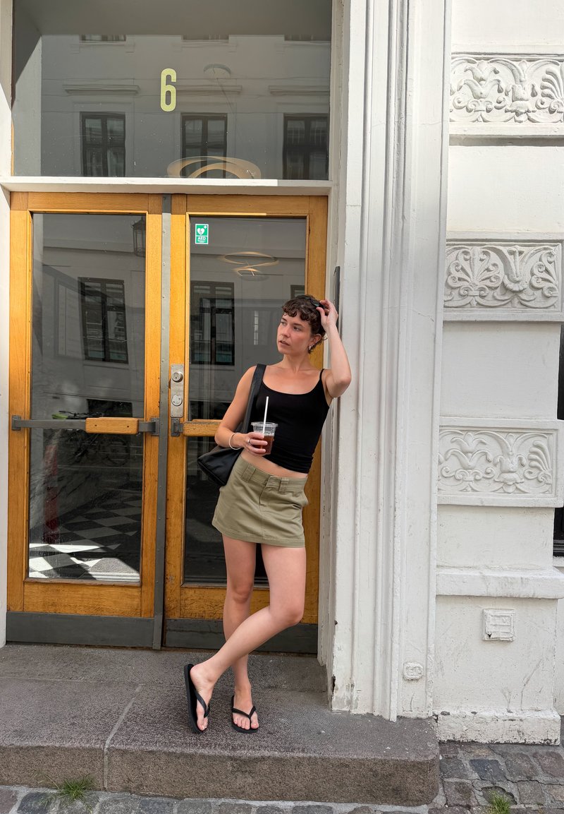 Jeune femme en haut noir, jupe beige et tongs tenant un verre, debout et appuyée contre l'encadrement orné blanc d'un bâtiment.