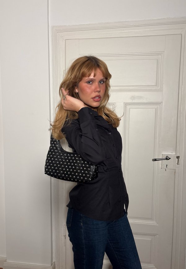 Jeune femme aux cheveux blonds mi-longs portant une chemise noire à boutons et un jean, tenant un sac à main noir clouté sur l’épaule.
