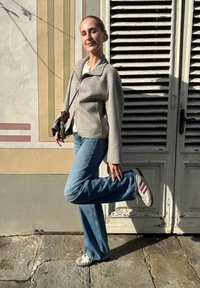 Γκρι μάλλινο blazer με κουδούνια, ανοιχτόχρωμο τοπ, μπλε φαρδιά τζιν και γκρι αθλητικά με κόκκινες ρίγες, ποζά�ροντας μπροστά σε μια υφασμάτινη τοιχογραφία.