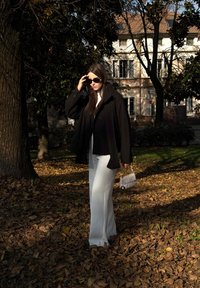 Palton negru, pantaloni largi albi și ochelari de soare negri. Ține o geantă mică albă. Scena include frunze căzute și un fundal cu clădiri.