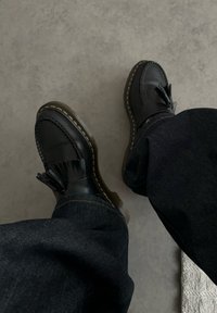 Fekete bőrcipők díszes bojttal, sárga varrással és sima felülettel. Sötét farmernadrággal párosítva, lazább megjelenést kölcsönözve.