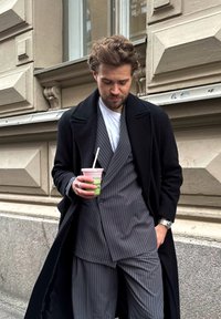 Homem em um sobretudo preto, terno cinza riscado, segurando um smoothie, com cabelo texturizado, de pé em frente a um prédio com detalhes clássicos.