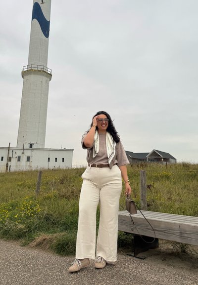 Una mujer está de pie en un área de césped, vistiendo pantalones de pierna ancha beige, una blusa marrón y una bufanda con estampado, sosteniendo un pequeño bolso. En el fondo se ve un faro alto.