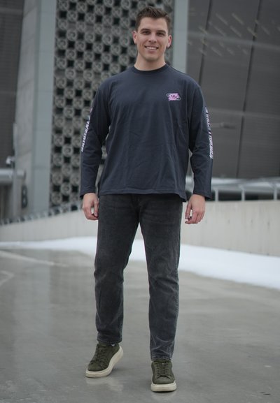 Joven de pie al aire libre sobre pavimento mojado, vistiendo camisa de manga larga oscura, jeans negros y zapatillas verde oliva, sonriendo a la cámara.