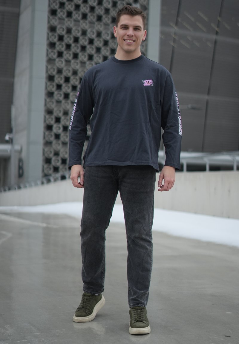 Jeune homme debout en plein air sur un trottoir mouillé, portant une chemise à manches longues foncée, un jean noir et des baskets vert olive, souriant à la caméra.