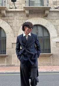 Dviejų sagų tamsiai mėlynas dryžuotas švarkas su plačiomis atlapomis, baltu apykakle ir dryžuota kakliene; derinamas su laisvomis, tamsiomis kelnėmis. Dėvimi dideli akiniai nuo saulės.