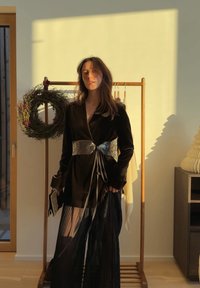Femme en tenue en velours noir avec ceinture métallique, debout à l'intérieur près d'un portant en bois avec une couronne et des vêtements suspendus.