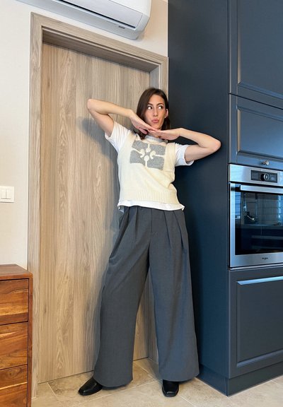 Chaleco blanco de punto con un diseño gris sobre una camiseta blanca, combinado con pantalones anchos gris oscuro y zapatos negros. De pie junto a una puerta de madera.