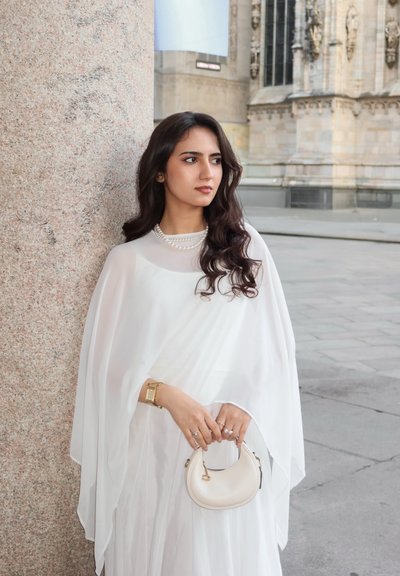 Mujer con vestido blanco y collar de perlas, de pie junto a una columna de piedra, sosteniendo un pequeño bolso beige, con un edificio histórico de fondo.