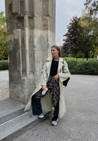 Světle béžový trench coat přes černý top, doplněný černými širokými džínami a kostkovanou košilí uvázanou v pase. Šedé tenisky a hrnek s kávou.