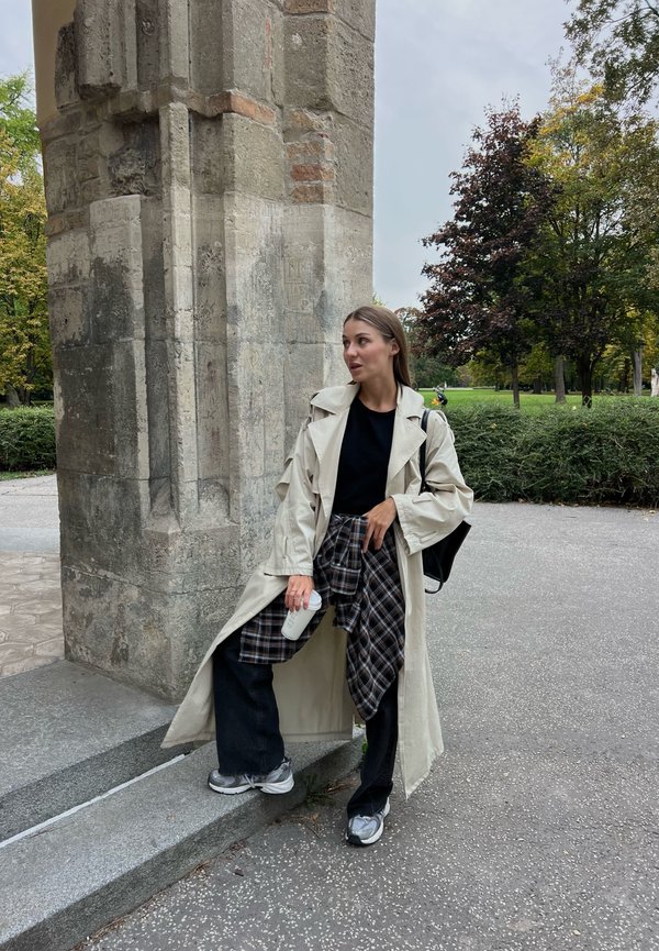 Ljusbeige trenchcoat över en svart topp, tillsammans med svarta vida jeans och en rutigt skjorta knuten i midjan. Grå sneakers och en kaffekopp.
