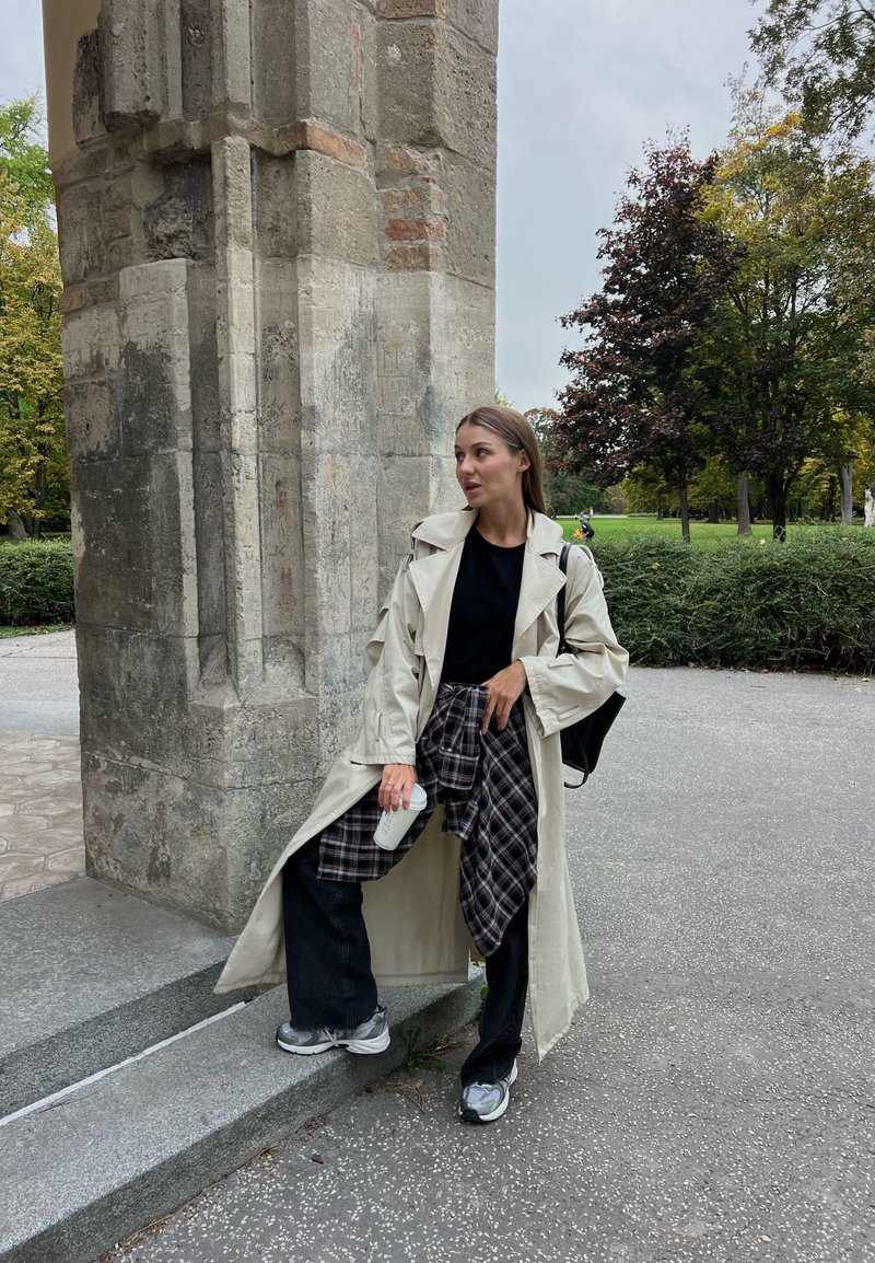Lichtbeige trenchcoat over een zwart top, gecombineerd met zwarte wijde jeans en een geruite blouse die om de taille is geknoopt. Grijze sneakers en een koffiekopje.