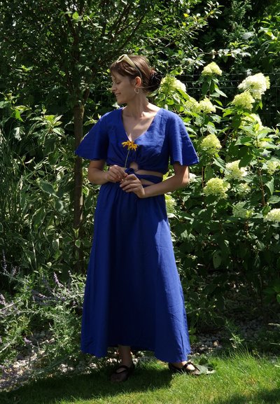 Vestido largo de lino azul con mangas cortas de volantes y un detalle de corte en la cintura, combinado con sandalias negras. Follaje verde en el fondo.