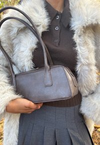 Persoon draagt een witte nepbonten jas, een bruin vest met knopen en een grijze geplooide rok, en houdt een kleine bruine leren handtas vast.
