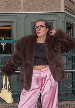 Chaqueta extragrande de piel sintética marrón, top negro corto, pantalones anchos de satén rosa y un pequeño bolso amarillo. Gafas y clips para el cabello complementan el look.