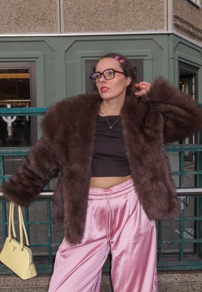 Chaqueta extragrande de piel sintética marrón, top negro corto, pantalones anchos de satén rosa y un pequeño bolso amarillo. Gafas y clips para el cabello complementan el look.