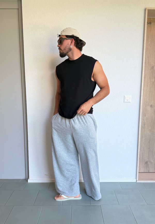 Hombre con una camiseta negra sin mangas, pantalones anchos grises, chanclas blancas, gafas de sol, pendientes y una gorra beige, de pie junto a una pared.