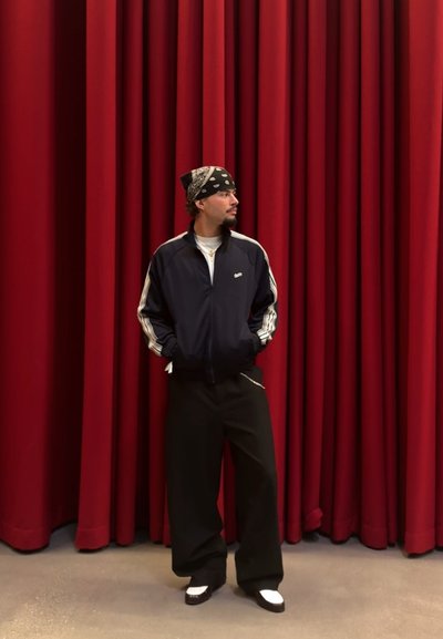 Chaqueta de chándal navy con rayas blancas, pantalones de pierna ancha negros y zapatos en blanco y negro, contra un fondo de cortina roja.