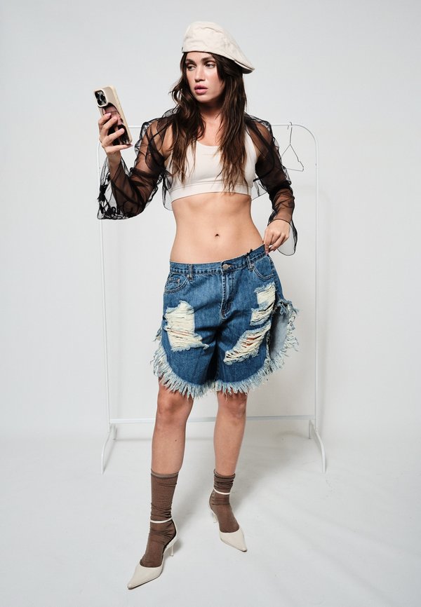 Frau trägt ein weißes bauchfreies Oberteil, transparente schwarze Ärmel, übergroße zerrissene Jeansshorts, beige Baskenmütze, hält ein Smartphone, richtet den Bund der Shorts.