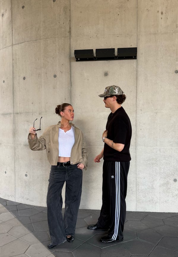 Deux jeunes adultes se font face devant un mur en béton, l’un tenant des lunettes de soleil, habillés de manière décontractée en vêtements de rue modernes.