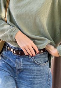 Cinto de pele castanho com ferragens prateadas, usado sobre calças de ganga de cintura alta, de denim azul claro, combinado com uma camisola verde de manga longa canelada.