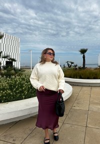 Πλεκτό κρεμ πουλόβερ σε chunky σχέδιο με διαμαντένιο μοτίβο, μπορντό σατέν φούστα, μαύρη τσάντα και γυαλιά ηλίου; εξωτερικός χώρος με φοίνικες στο παρασκήνιο.