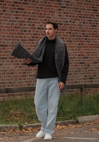 Grauer und schwarzer karierter Schal, gehalten von einer Person in einem schwarzen Pullover, hellblauen Jeans und weißen Sneakers, vor einem Ziegelwand-Hintergrund.