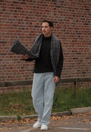 Bufanda a cuadros gris y negra sostenida por una persona que lleva un suéter negro, jeans azul claro y zapatillas blancas, contra un fondo de pared de ladrillo.