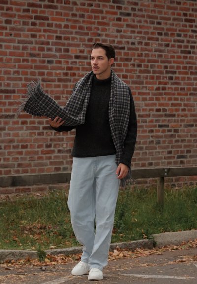 Bufanda a cuadros gris y negra sostenida por una persona que lleva un suéter negro, jeans azul claro y zapatillas blancas, contra un fondo de pared de ladrillo.