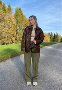 Ruda odinė striukė su aukščiu atlenkiamu apykakle, po ja žalia marškiniai, šviesiai rusva palaidinė ir alyvuogių žalumo kelnės, užbaigta šviesiais sportbačiais.