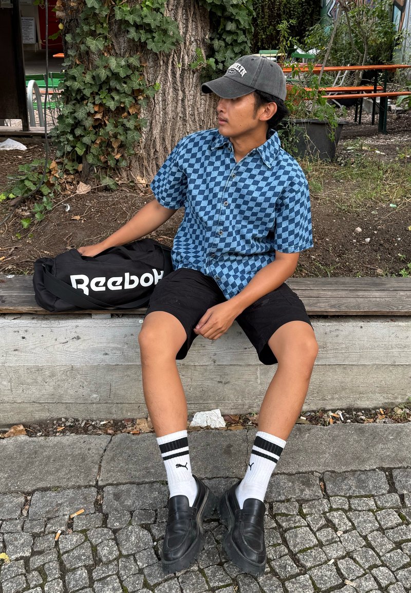 Persona con camicia a quadri a maniche corte, pantaloni corti neri e cappello è seduta su una panchina all'aperto, tiene in mano una borsa Reebok nera, vicino a un albero e del verde.