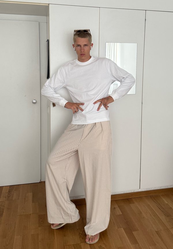 Personne portant une chemise blanche à manches longues, un pantalon beige à larges rayures, des tongs, des lunettes de soleil sur la tête et des écouteurs, debout à l'intérieur avec les mains sur les hanches.
