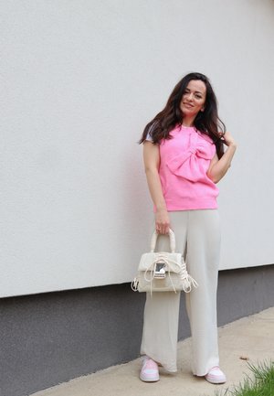 Un suéter sin mangas rosa con un gran lazo, combinado con pantalones de pierna ancha beige claro y zapatillas rosas. Lleva un bolso blanco texturizado.