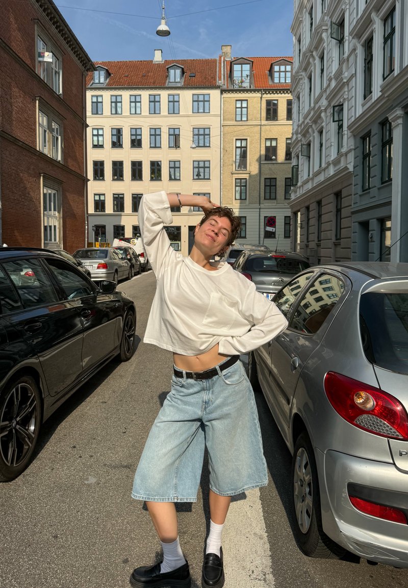 Camisola branca de mangas compridas, cropped, culottes de ganga azul claro, sapatos pretos e meias brancas. Cenário urbano com carros estacionados e edifícios.