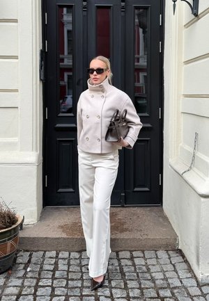Mujer con chaqueta cruzada color crema y pantalones blancos, sosteniendo un bolso oscuro, de pie frente a una puerta negra con suelo de adoquines.