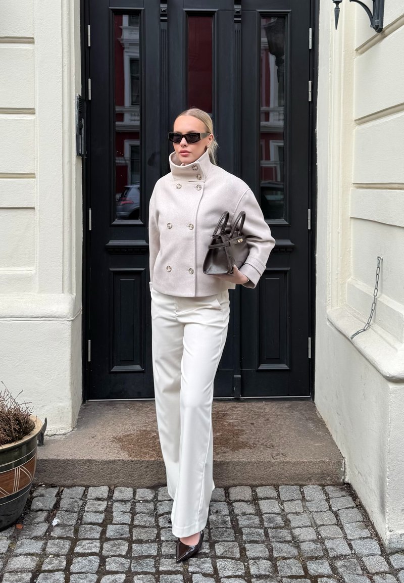 Femme portant une veste croisée crème et un pantalon blanc, tenant un sac à main foncé, debout devant une porte noire sur un sol pavé.